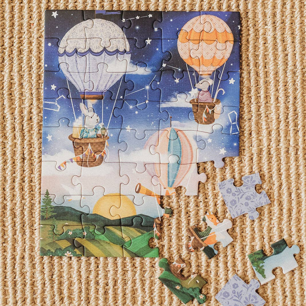 A partially completed Sky High Friends 40 Piece Mini puzzle, featuring whimsical animals in hot air balloons floating over a sunrise landscape, lies on a textured carpet with a few loose pieces beside it.