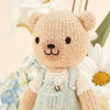 Cuddle + Kind Tiny Charlie the Honey Bear, a hand-knit doll in light blue overalls with a bee detail, stands before white daisies and light blue hydrangeas. Handmade in Peru.