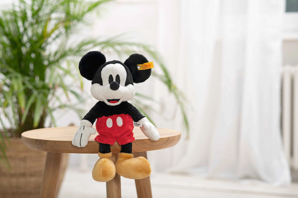 A Steiff, Disney's Mickey Mouse Stuffed Plush Toy, 12 Inches sits on a small wooden table. The cartoon mouse character, with black ears, red shorts, and yellow shoes, is set against a bright background featuring green potted plants and sheer white curtains.