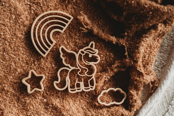 The Unicorn Cutter Set - Eco Playdough Tool features four eco-friendly cookie cutters shaped like a unicorn, rainbow, star, and cloud arranged on a textured brown fabric surface—ideal for sensory play. The unicorn playdough cutter brings a magical element to creative activities.