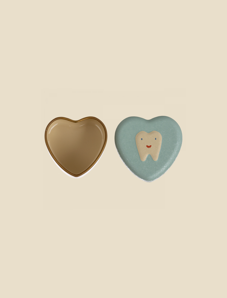 A heart-shaped box with a beige, reflective surface, open to reveal its beige interior, sits beside a blue lid decorated with a smiling cartoon tooth. This charming Maileg Tooth Box evokes childhood memories and serves as a perfect baby teeth box. The background is a neutral, light beige color.