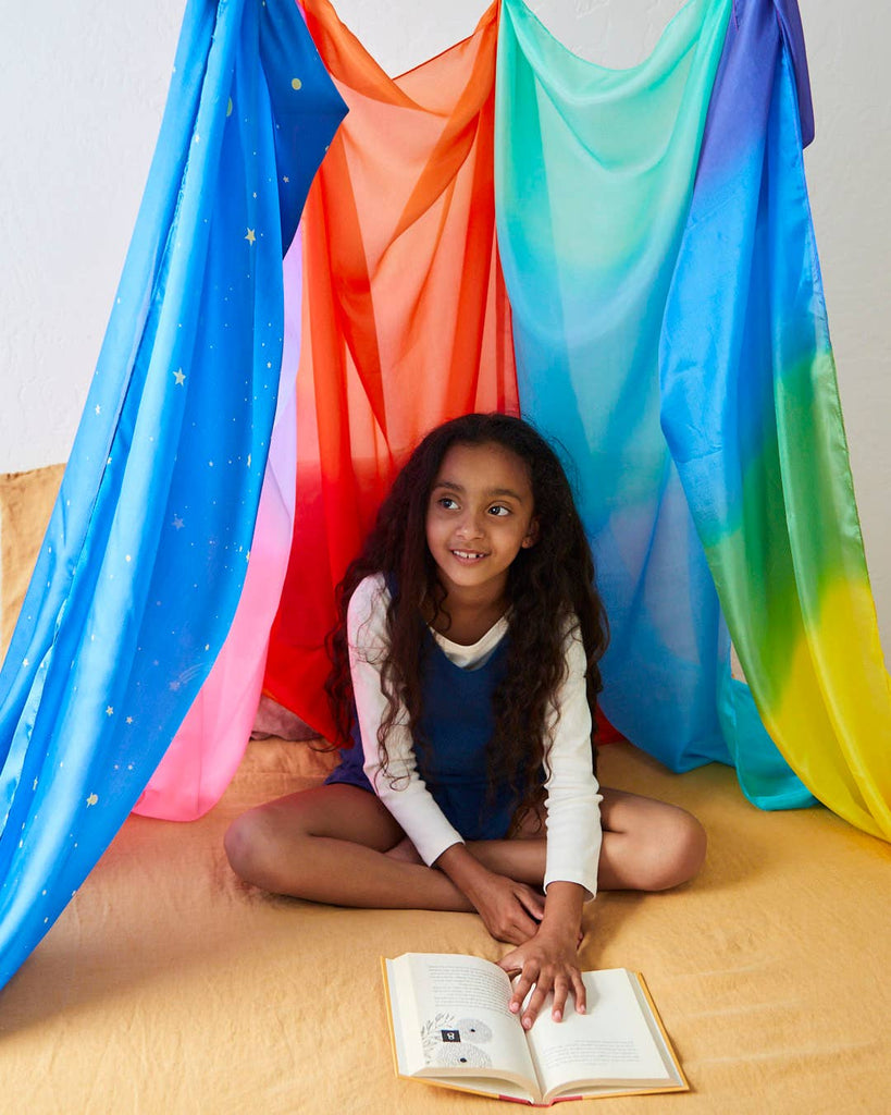 A child with long hair sits cross-legged on a yellow blanket, smiling and holding an open book. Sarah's Silk Giant Ocean Playsilk in shades of blue, red, orange, and green drapes around them, forming a vibrant tent-like structure that inspires open-ended play in an imaginative world.