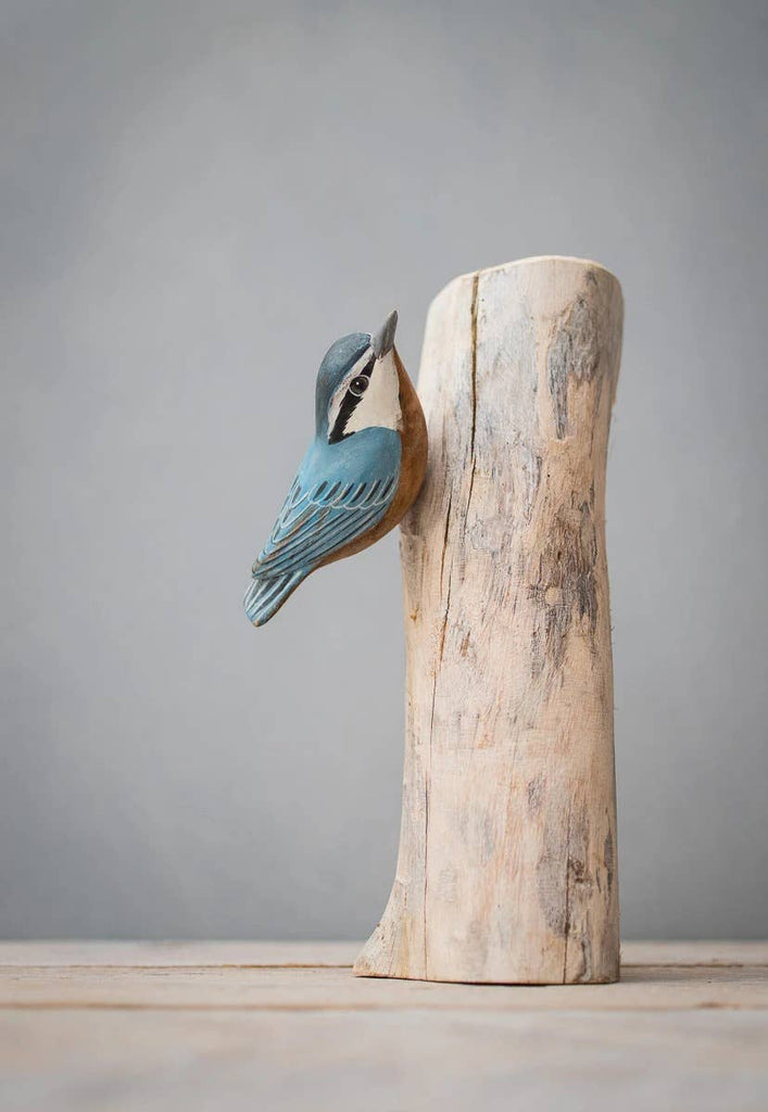 The Hand Carved Ornament - Red Breasted Nuthatch Bird- 8"H shows a wooden bird clinging to a smooth, softwood tree trunk, inspired by the Red Breasted Nuthatch, set against a plain gray background.