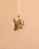 The Bukowski - Kharma Dog Bag Charm, a small plush dog keychain with curly brown fur, hangs from a beige ribbon against a plain light brown background.