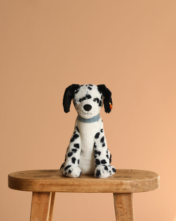 The Steiff Oskar Dalmatian Dog plush, 11 inches tall with black spots and floppy ears, sits on a wooden stool against a plain beige background.