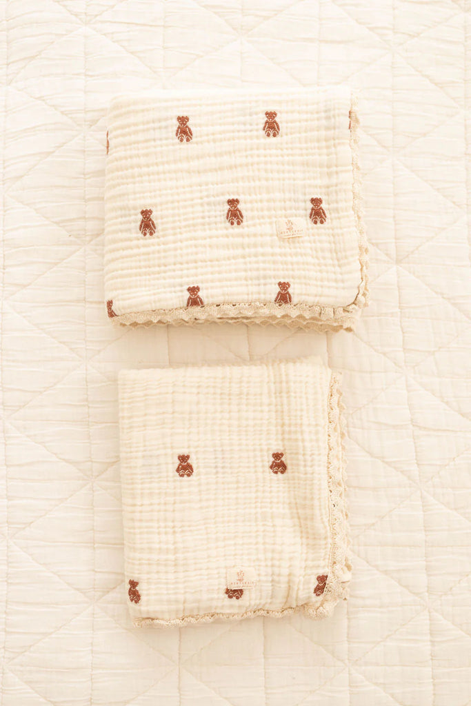 Two Organic Cotton Layered Gauze Baby Blankets - Teddy (ships in one week), cream-colored with small brown teddy bear patterns and lace-trimmed edges, are neatly folded side by side on a quilted white surface.