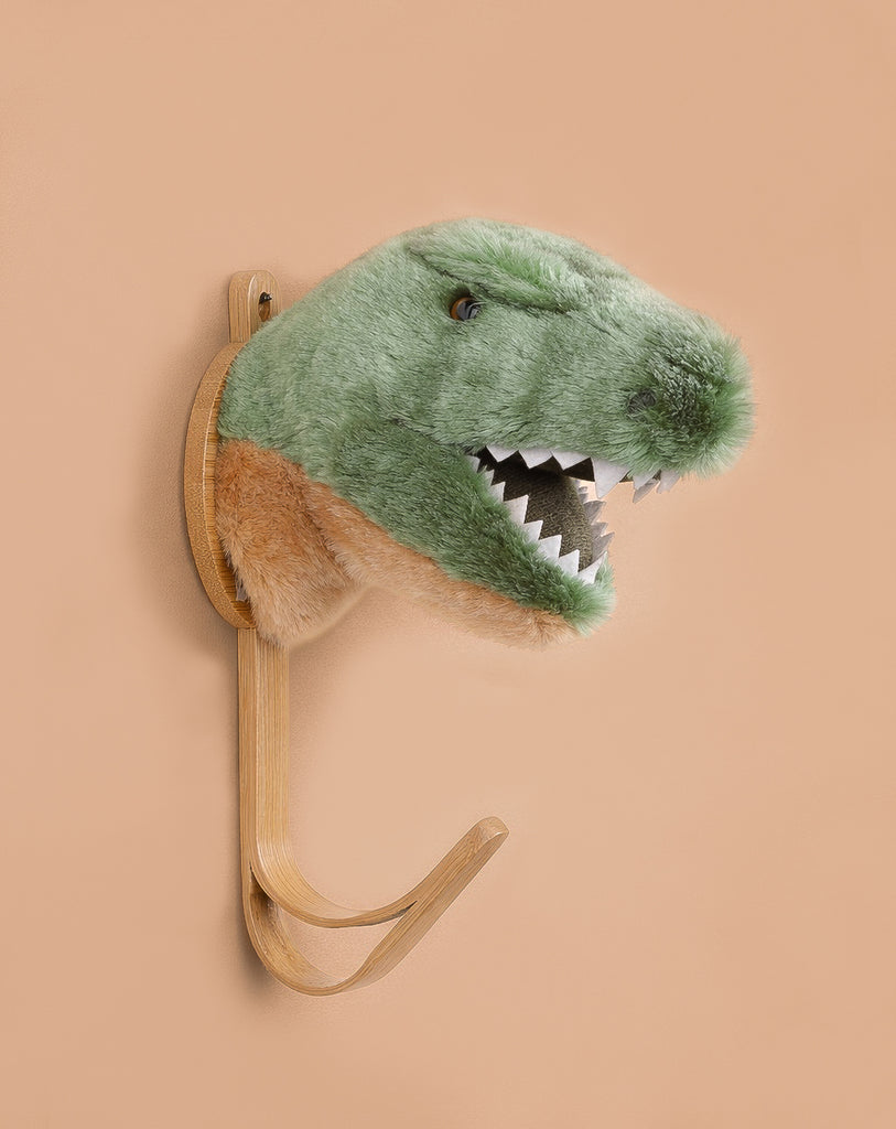 The Coat Hanger - T-Rex Dino is a wall-mounted hook with a plush green and beige dinosaur head featuring felt teeth, designed to resemble a T. rex, displayed against a plain beige background.