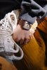 A person wearing a gray sweatshirt and mustard yellow pants holds a white woven bag. On their left wrist, they sport the Mini Kyomo Watch - Plum Cake with a brown band.