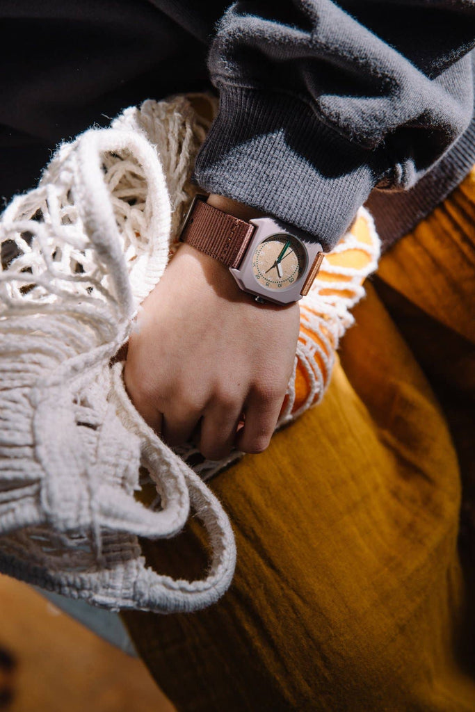 A person wearing a gray sweatshirt and mustard yellow pants holds a white woven bag. On their left wrist, they sport the Mini Kyomo Watch - Plum Cake with a brown band.