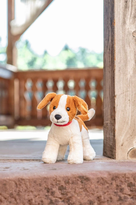 A Steiff Snuffy Dog plush toy with white and brown fur, sitting on a stone surface, with a blurred wooden fence in the background.