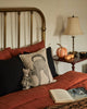 A cozy bed with rust-colored bedding and black pillows has a cushion with a black cat, a small mouse doll, and the Coral & Tusk Bat Surprise Doll. An open book sits on the bed. A side table displays a lamp and decorative pumpkins.