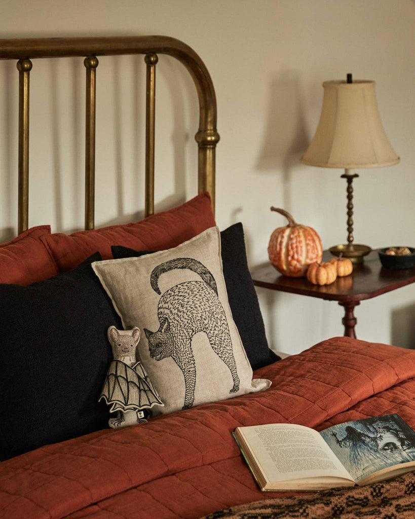 A cozy bed with rust-colored bedding and black pillows has a cushion with a black cat, a small mouse doll, and the Coral & Tusk Bat Surprise Doll. An open book sits on the bed. A side table displays a lamp and decorative pumpkins.