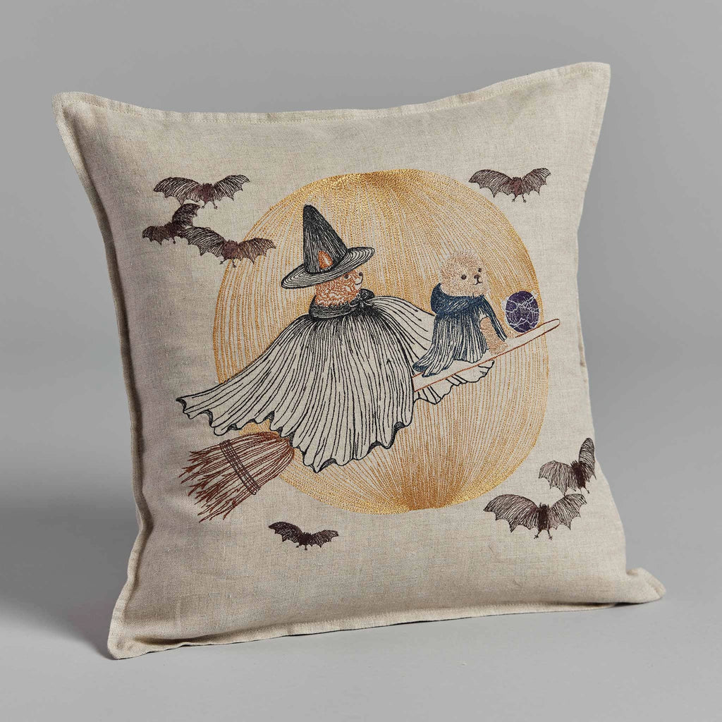 The Coral & Tusk Supermoon Spell Pillow showcases a whimsical Halloween illustration of a witch in a gray cape and hat, soaring on her broomstick with a small cat companion. They are set against an orange moon backdrop, encircled by several flying bats, all on a light beige background.