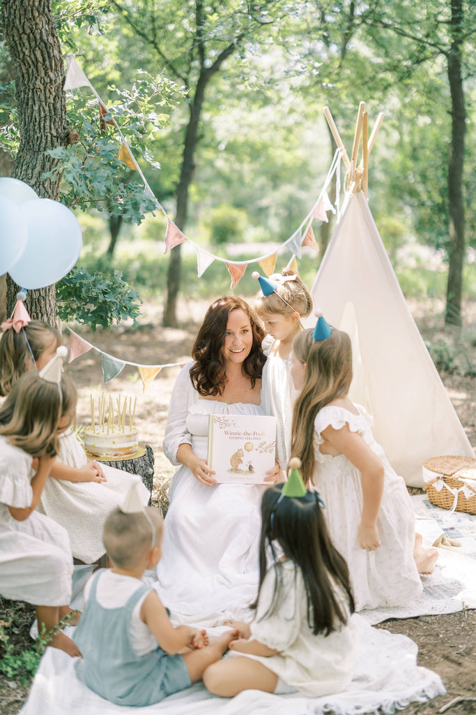 A woman reads Winnie-the-Pooh: Finding Feelings to kids at a picnic in the woods, surrounded by balloons and banners that create a festive outdoor vibe.