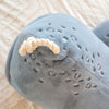 A detailed close-up of the Ride-On Whale Pouf shows its soft blue plush body with textured spots, knotted rope tail, and stitched accents, resting on patterned white fabric and capturing a playful whale-inspired charm.