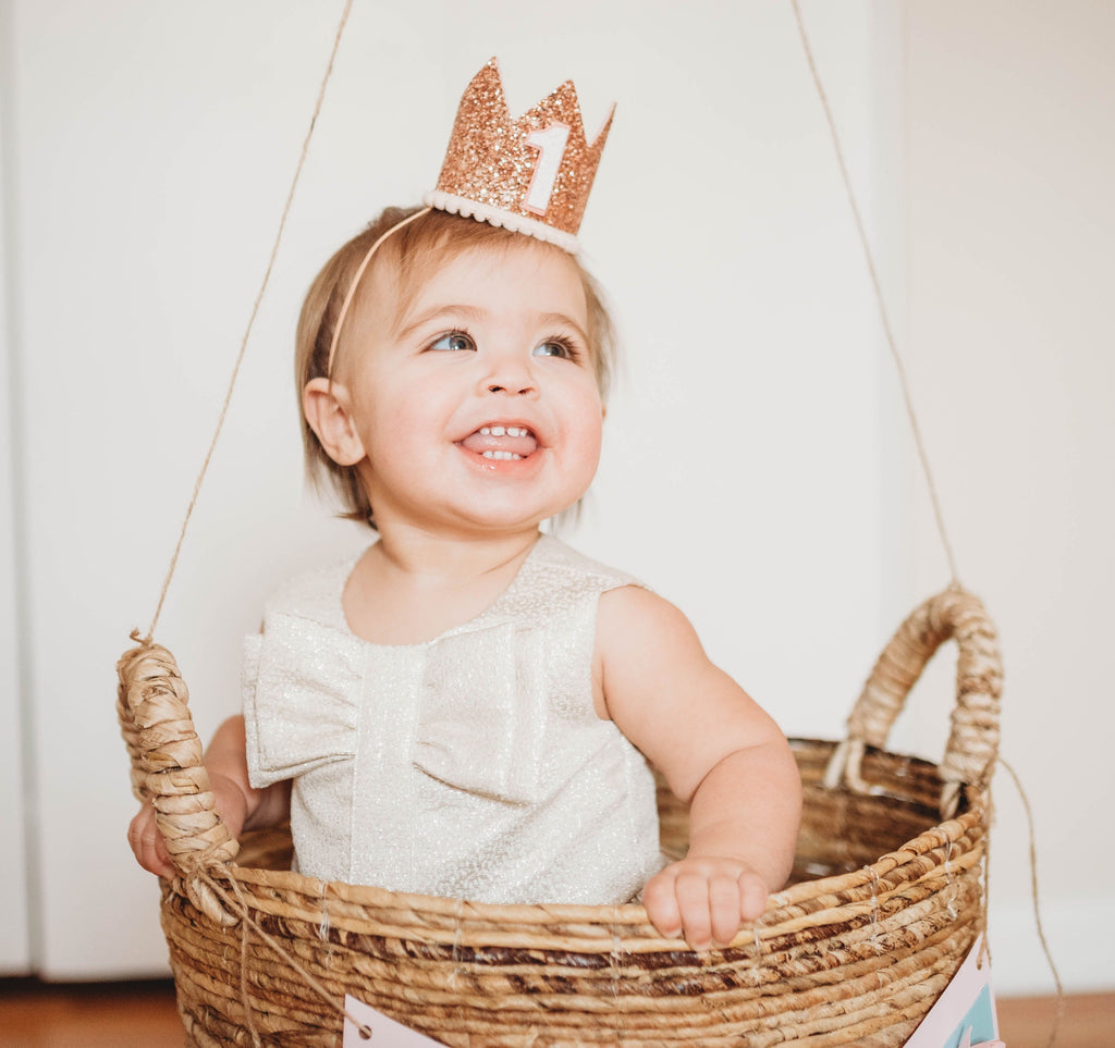 A smiling toddler with a "1" Rose Gold Glitter Crown sits inside a woven basket. The child is wearing a light-colored dress with a large bow on the front. The basket, perfect for birthday celebrations, hangs from ropes and is adorned with a string of small colorful flags.