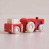The No. 34 GT190 Tilly, a vintage red wooden tractor with a matching trailer carrying a wooden hay bale and featuring light brown wheels, sits on a smooth light pink surface—an inviting farmyard toy made in England, perfect for imaginative play.