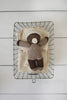 A brown and beige Hand-Knit Baby Doll - Cocoa lies snugly wrapped in an organic cotton swaddle within a rectangular wire basket, placed on a white wooden surface.