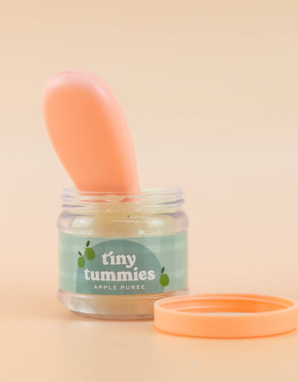 A small jar of Tiny Tummies Apple Jelly Food, featuring a peach-colored lid from Tiny Harlow, is open on a surface. A magic spoon stands upright in the jar, ready for little ones to enjoy. The background is a soft beige.