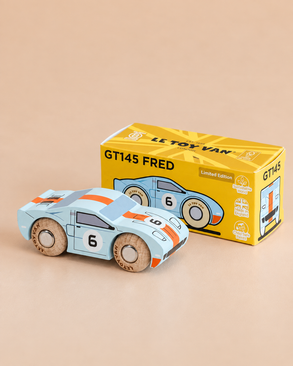 A blue classic toy car with the number 6 sits next to its yellow box labeled “No. 26 GT145 Fred | Made in England” on a beige surface. The box features an illustration of the wooden racing car and some icons.