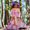A young child in a forest wears Sarah's Silks Magical Creatures Playsilk - Pink Unicorn as a petal-patterned cape and a purple headband with large ears, smiling while sunlight filters through the tall trees behind her.