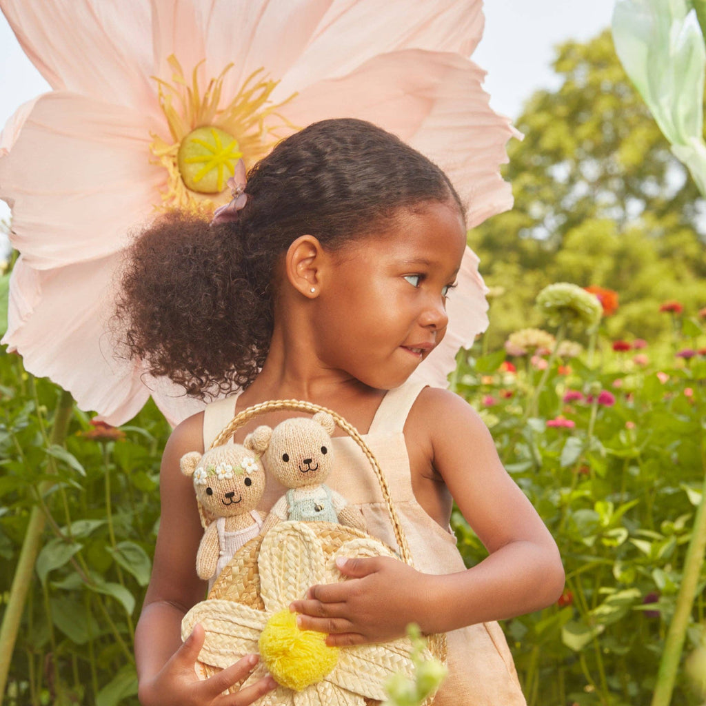 A young girl in a light dress stands in a garden, holding a basket with two teddy bears, including the Cuddle + Kind Tiny Charlie the Honey Bear. She is surrounded by large flowers and greenery, smiling as she looks to her right.