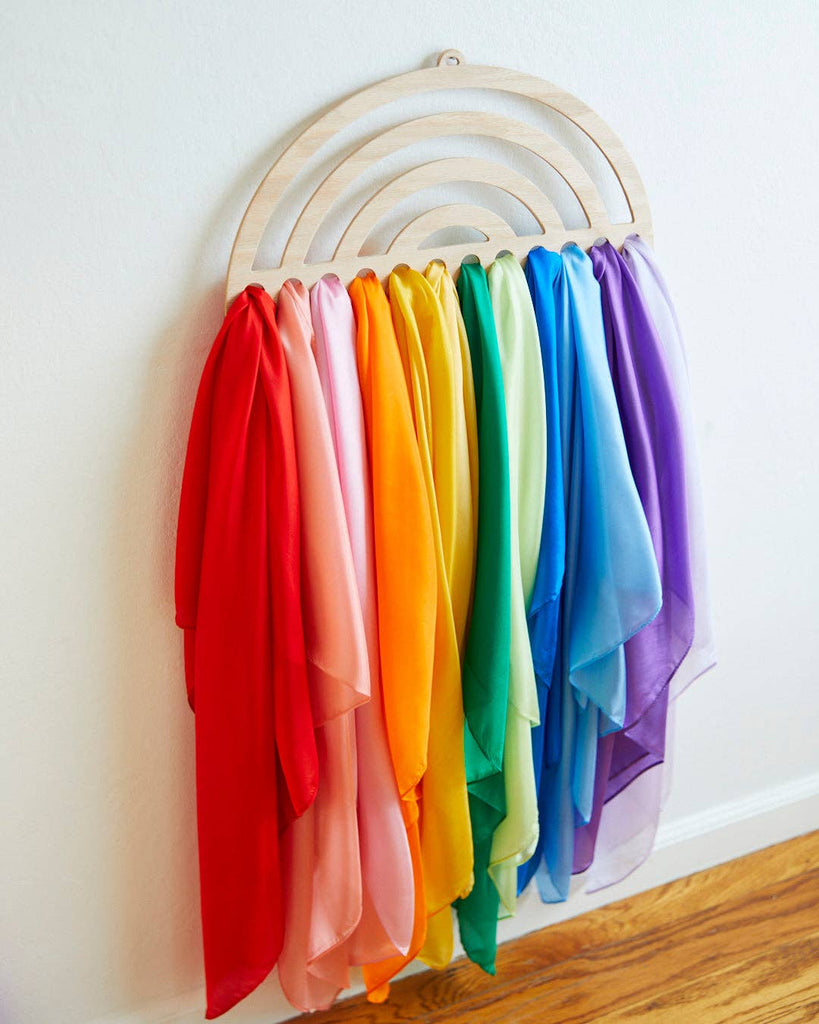 In a vibrant playroom, Sarah's Silk Large Playsilk Display - Rainbow, beautifully crafted from Red Oak MDF in a rainbow shape, adorns the white wall. Elegantly draped over the rack are colorful playsilk pieces in shades of red, pink, orange, yellow, green, blue, and purple that cascade towards the wooden floor.