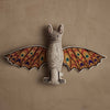 The Coral & Tusk Bat Surprise Doll, featuring colorful patterned wings in red, orange, green, and blue, hangs on a textured beige wall.