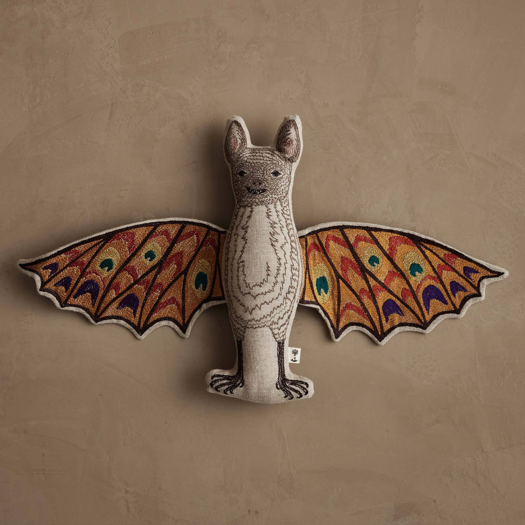 The Coral & Tusk Bat Surprise Doll, featuring colorful patterned wings in red, orange, green, and blue, hangs on a textured beige wall.