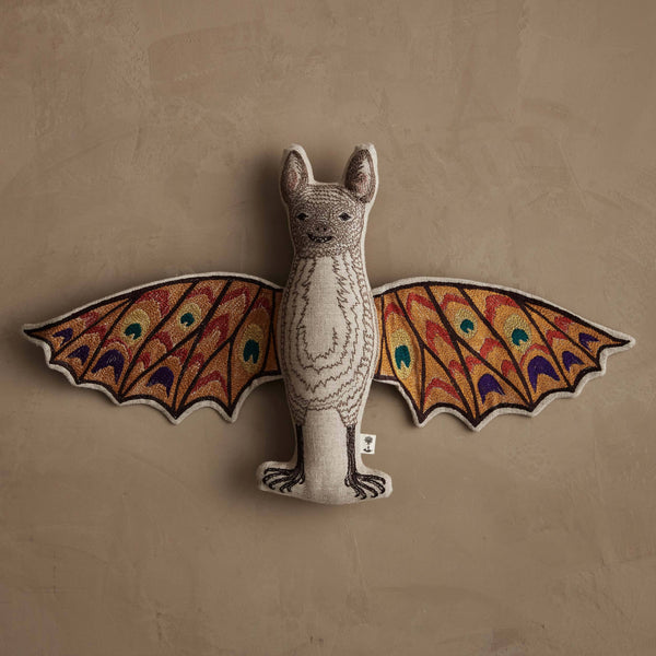 The Coral & Tusk Bat Surprise Doll, featuring colorful patterned wings in red, orange, green, and blue, hangs on a textured beige wall.