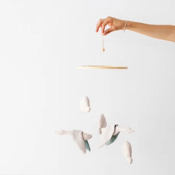A hand holding a Cloth Clouds and Birds Mobile with four fabric birds crafted from organic cotton muslin in white and grey hues, floating against a clean white background.