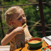 A young girl in a yellow plaid dress sits at a wooden table outdoors, eating soup from the Wally the Watermelon Bowl & Spoon Set. Greenery and a railing are visible in the background.