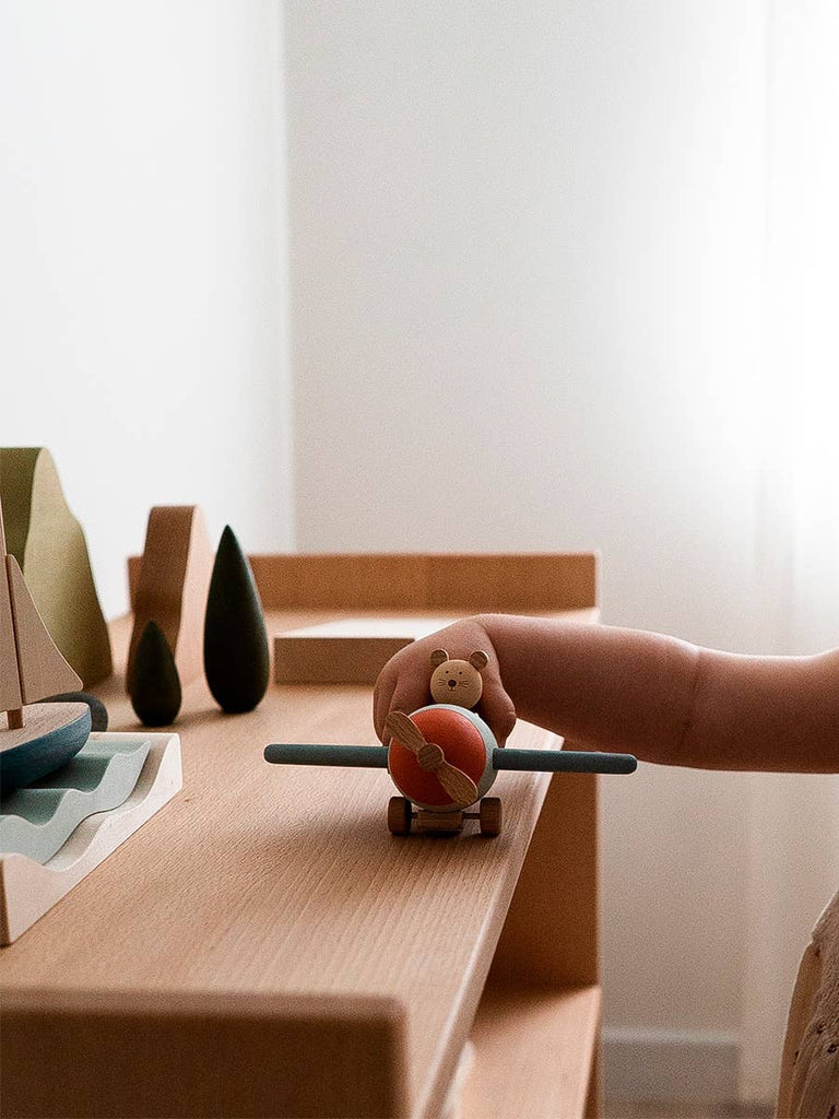 A child’s hand holds the Sabo Concept Aviator Mouse on a wooden table, surrounded by minimalist wooden trees—an inviting scene for imaginative play in a softly lit room.