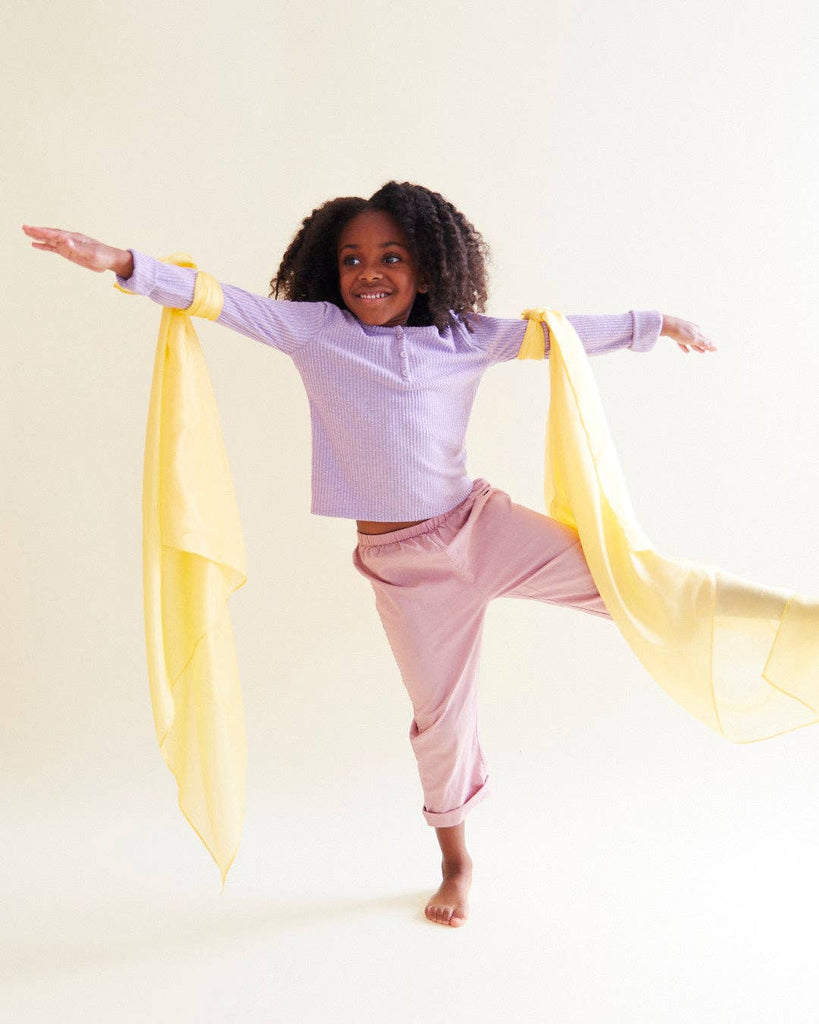 A young child with curly hair balances on one leg, arms outstretched, smiling as colorful Sarah's Silks Solid Color Playsilk - Sunshine is tied to each arm—perfect for open-ended play against a plain light background.