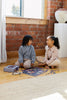 Two children sit on a wooden floor by a brick wall, smiling at each other as they work together on the Courting Critters 500 Piece Puzzle. Sunlight streams through a nearby window, creating a warm and inviting atmosphere.