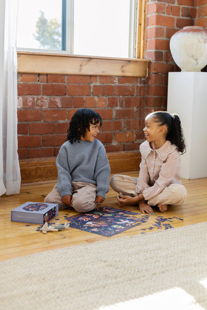 Two children sit on a wooden floor by a brick wall, smiling at each other as they work together on the Courting Critters 500 Piece Puzzle. Sunlight streams through a nearby window, creating a warm and inviting atmosphere.