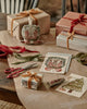 A gift wrapping scene with presents, ribbon, scissors, and greeting cards on a wooden table. Featured: Coral & Tusk Peek a Tree Card with woodland and holiday designs like a fox and Christmas tree.