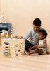 Two children sit on a light wooden floor, playing with the Wooden Activity Cube. The older child guides the younger as they explore beads and interactive features that support fine motor skill development.