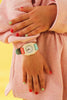 A person in a pink outfit displays the Mini Kyomo Watch - Bloom, featuring a turquoise and pink strap. Their red and mint green nails stand out against the vibrant yellow background.