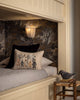 A cozy bed with an Oeko-Tex certified Coral & Tusk Full Moon Masquerade Pillow featuring forest creatures, set against dark floral wallpaper. A wooden stool with a bird figurine and a warm wall light complete the scene.