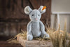 A Steiff Cheesy Mouse Stuffed Animal with large ears, a pink nose, and bean bag stuffing sits on hay-covered wood, featuring a yellow ear tag and wheat stalks softly blurred in the background.