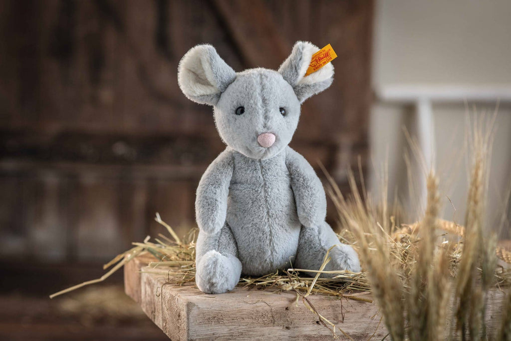 A Steiff Cheesy Mouse Stuffed Animal with large ears, a pink nose, and bean bag stuffing sits on hay-covered wood, featuring a yellow ear tag and wheat stalks softly blurred in the background.
