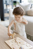 A young child in light overalls concentrates while placing a wooden animal piece from the “Farm Animals Wooden Puzzle: A Tabitha Paige Puzzle” into its spot on a wooden table in a bright, modern living room, exploring the fun of this toddler educational toy.
