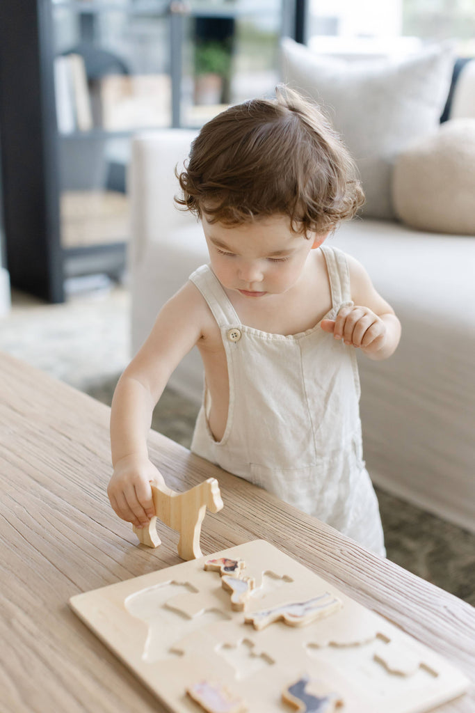 A young child in light overalls concentrates while placing a wooden animal piece from the “Farm Animals Wooden Puzzle: A Tabitha Paige Puzzle” into its spot on a wooden table in a bright, modern living room, exploring the fun of this toddler educational toy.
