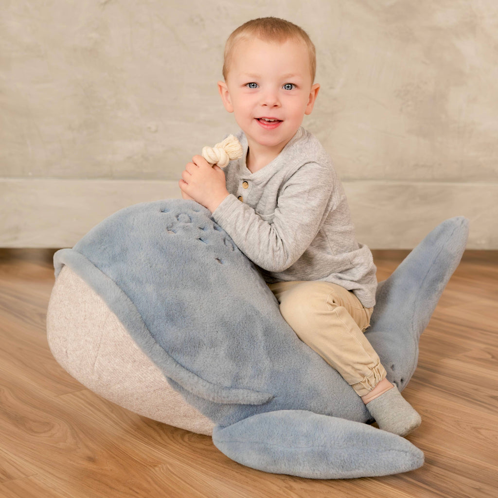 A young child with short blond hair, dressed in a gray shirt and beige pants, smiles while sitting on the Ride-On Whale Pouf on a wooden floor and holds a knotted rope toy.