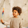 A young child with curly hair and a beige sweater gazes at a Grimm's Spirelli Clatter Wand held in their hand, its vibrant, cascading wooden rings capturing their curiosity. The background is softly blurred with string lights visible.