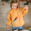 A young child wearing an orange hoodie holds the Grimm's Spirelli Clatter Wand, watching with focus as its colorful beads spiral in a rainbow pattern, reminiscent of the careful attention children give to cascading rings or a wooden spiral toy.