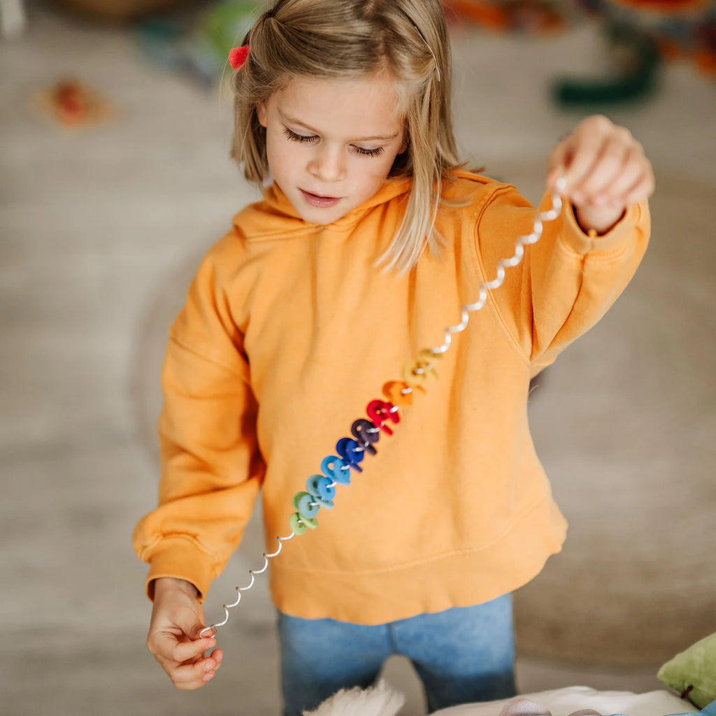 A young child wearing an orange hoodie holds the Grimm's Spirelli Clatter Wand, watching with focus as its colorful beads spiral in a rainbow pattern, reminiscent of the careful attention children give to cascading rings or a wooden spiral toy.