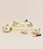 The eco-friendly Wooden Tea Set & Tray includes a floral-designed teapot, two cups, saucers, a tray, two heart-shaped biscuits, and a butter knife on a plain light beige background. Perfect for imaginative play, this charming set ships in approximately one week.