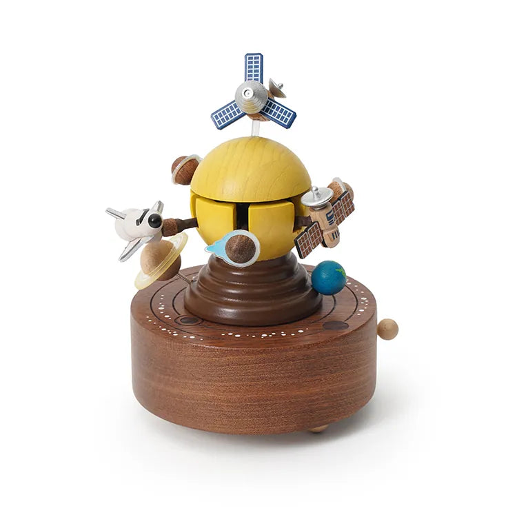 The Space Exploration Music Box by Wooderful Life is a hand-cranked music box featuring a yellow ringed planet, orbiting satellites, a rocket, and small planets on a round wooden base with white decorative markings.